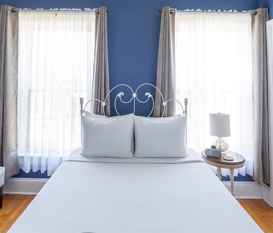 A neatly made bed with white pillows and a lamp beside it, framed by light curtains and a blue wall.