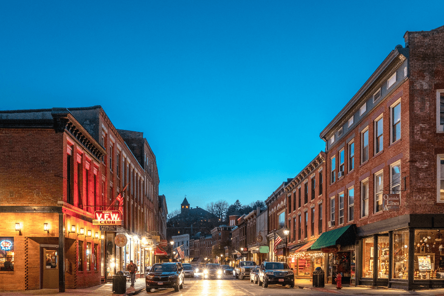 A vibrant street scene at dusk featuring historic brick buildings, shops, and cars along the roadway.